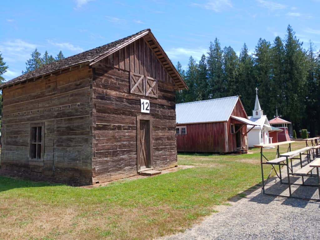 Row of 3 historic buildings at Berthusen Park Lynden WA. The closest is a log cabin, the next is an old red shed, and in the background is a small white chapel