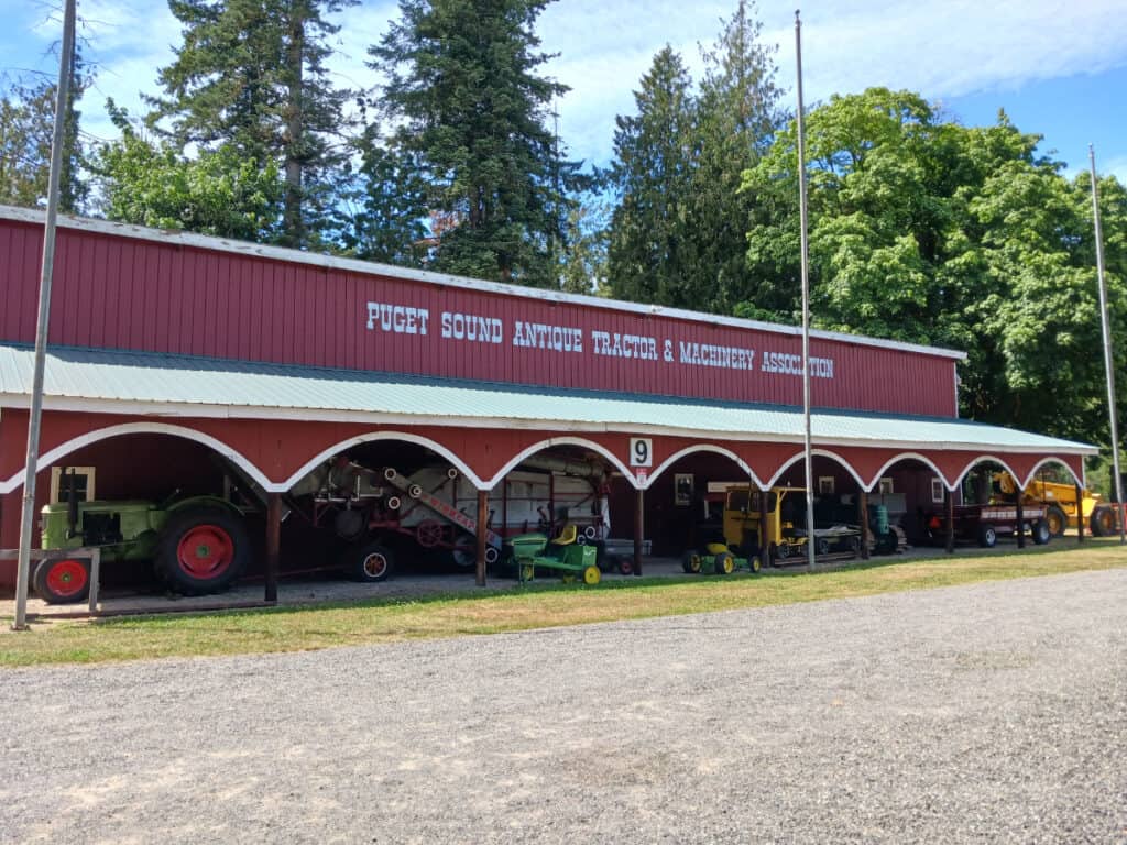 Red storage barn with text Puget Sound Antique Machinery Association with antique equipment under the eaves.