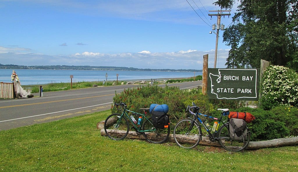 two bicycles parked outside sign for Birch bay state park. Grass in foreground and the bay and blue sky in the background