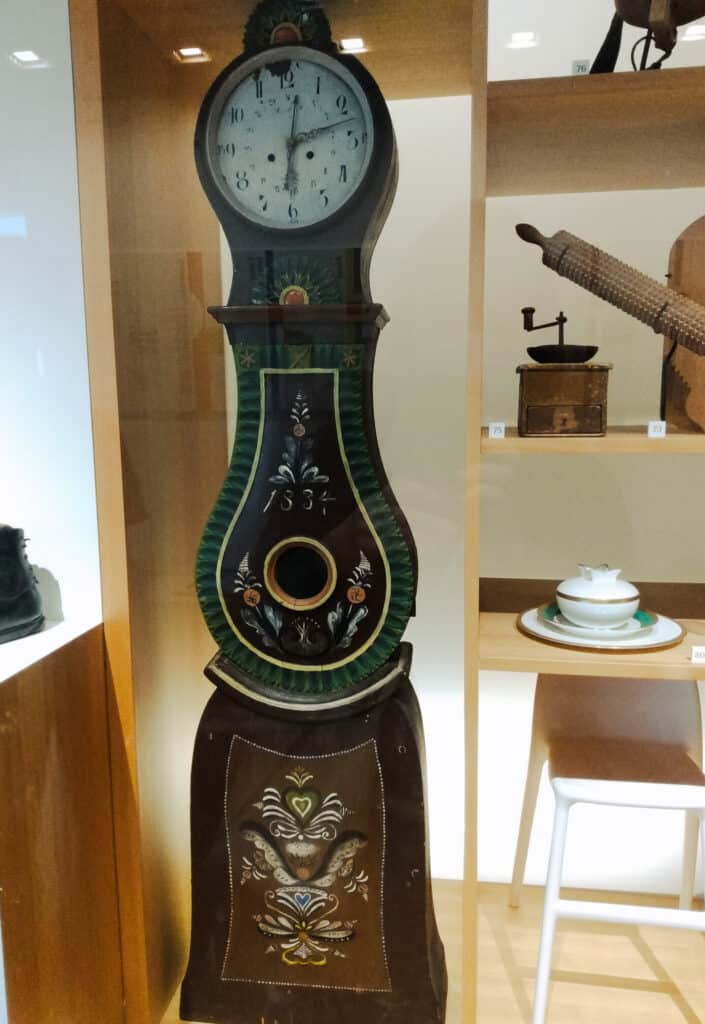 scandinavian grandfather clock with folk art painting on it, to the side of it are antique kitchen objects, nordic museum, ballard
