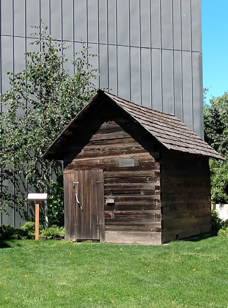 log hewn finnish style sauna, small cabin on grass, in background is side of nordic museum in ballard with gray exterior
