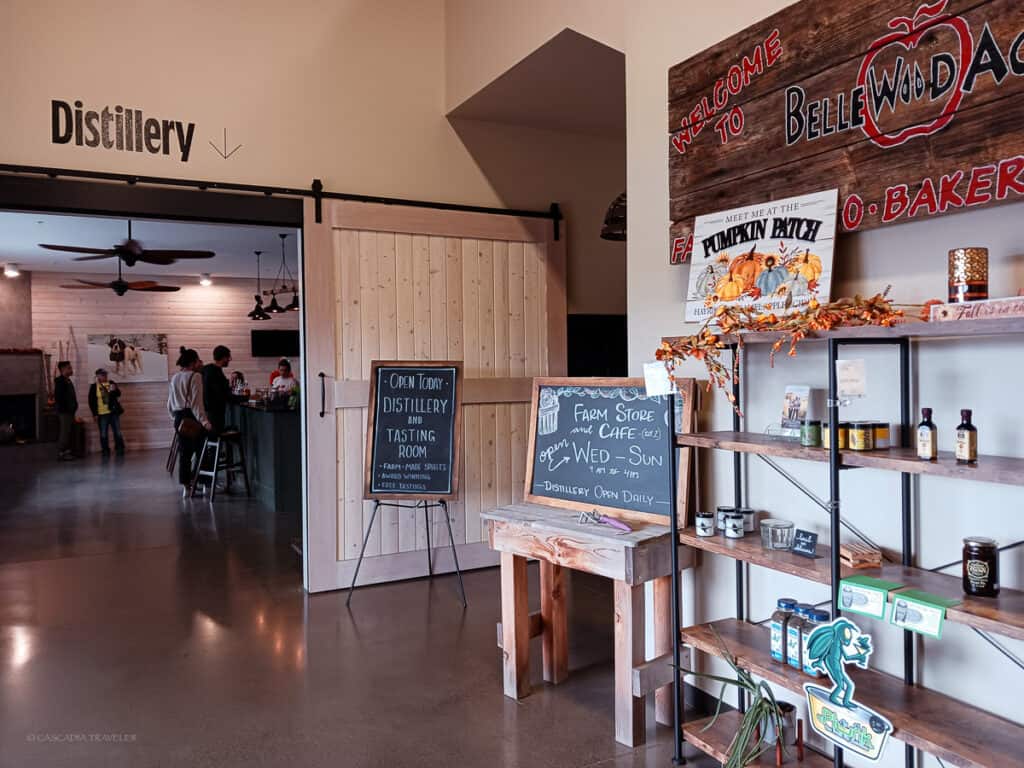 shelf with assorted bottle and jars at the Bellewood Acres farm store, wooden barn door leading to distillery tasting room. Chalkboard signage with opening hours.

