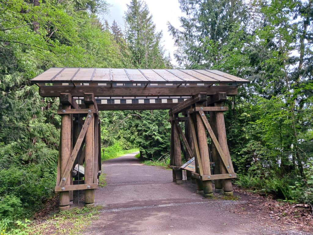 Wooden archway in the style of a railway trestle over a trail entrance. Large wide path with trees on either side.