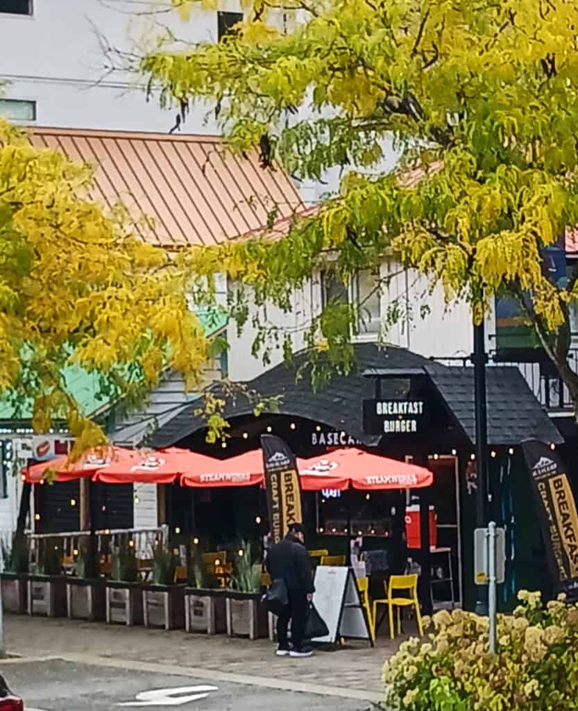 black shingled restaurant Basecamp Burger Joint in Harrison Hot Springs; in foreground are red umbrellas and outdoor seating along the sidewalk
