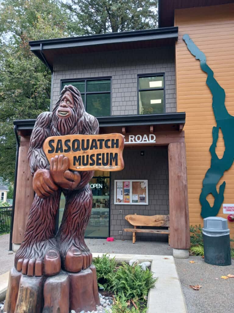 wooden statue of a sasquatch holding a sign that says sasquatch museum, behind is a contemporary building; Harrison Hot Springs Visitor Centre