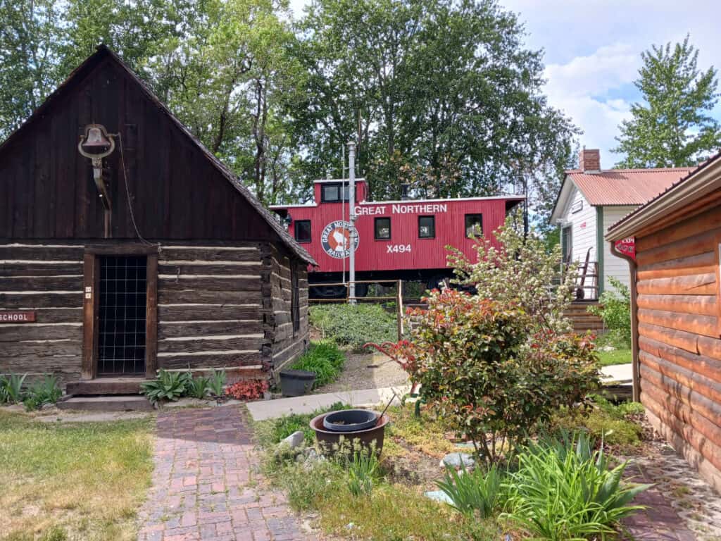 exterior shot at pioneer village Cashmere with schoolhouse cabin in foreground, bushes and grasses landscaping, Great Northern red caboose in background