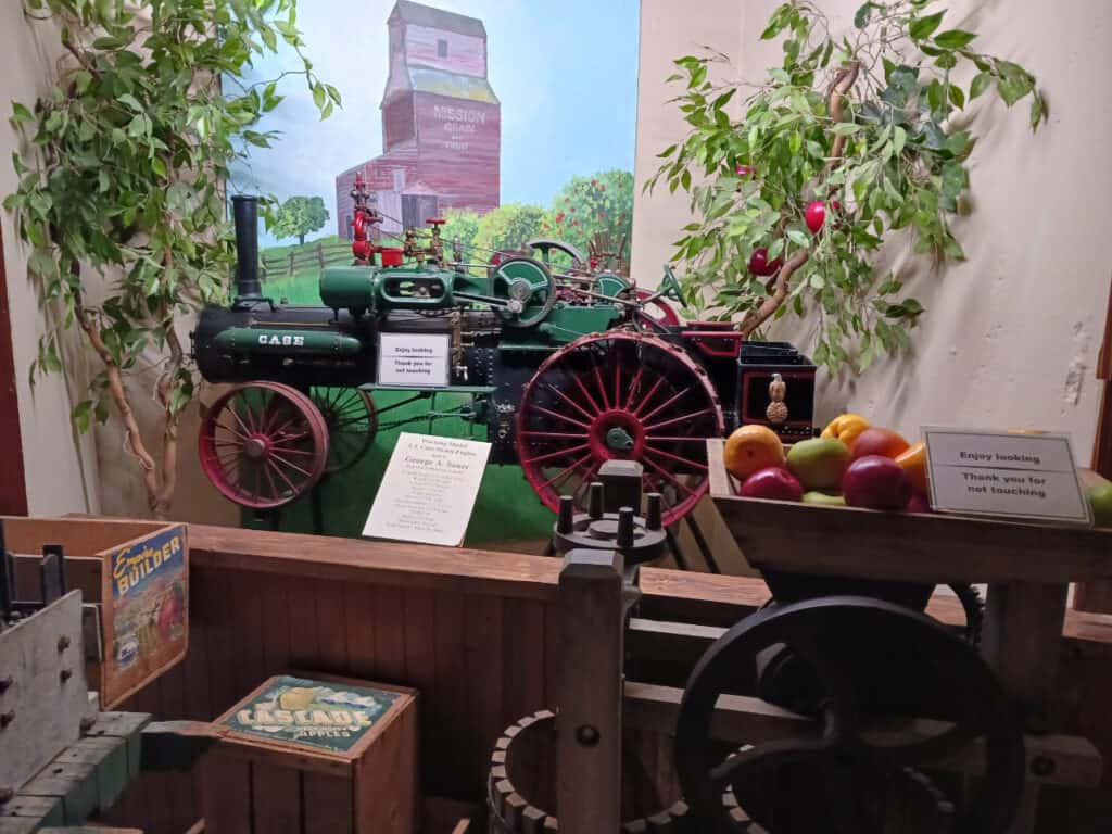 a museum display with an antique case tractor green with red wheels, painting of a grain elevator in background and fake apple tree,  fruit in foreground, at Cashmere museum