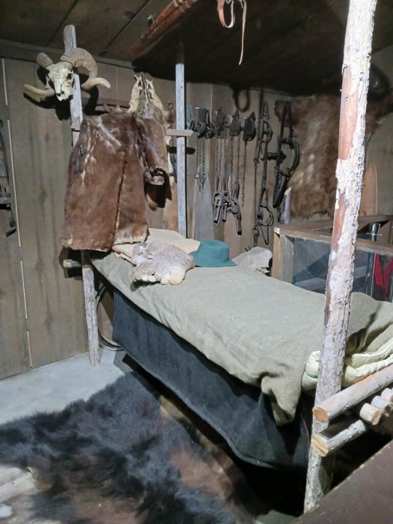 narrow bed with grey blanket, bedframe made of round fence posts, skins, furs and tools decorate the walls and floor, exhibit inside Cashmere museum